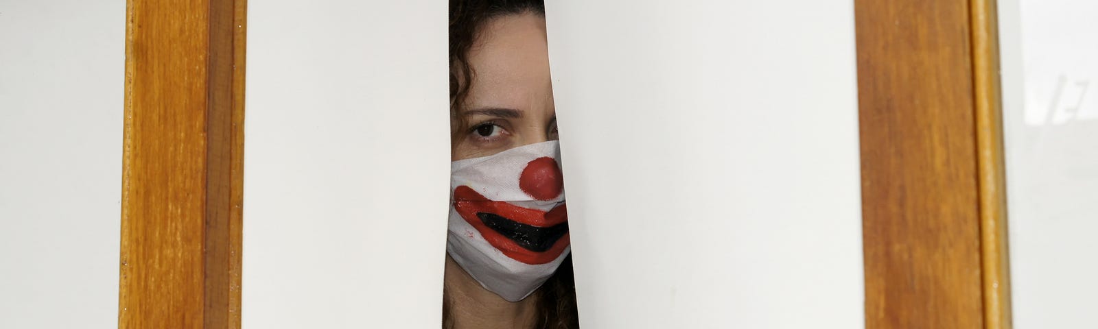 A woman wearing a Joker face mask peering through a crack.