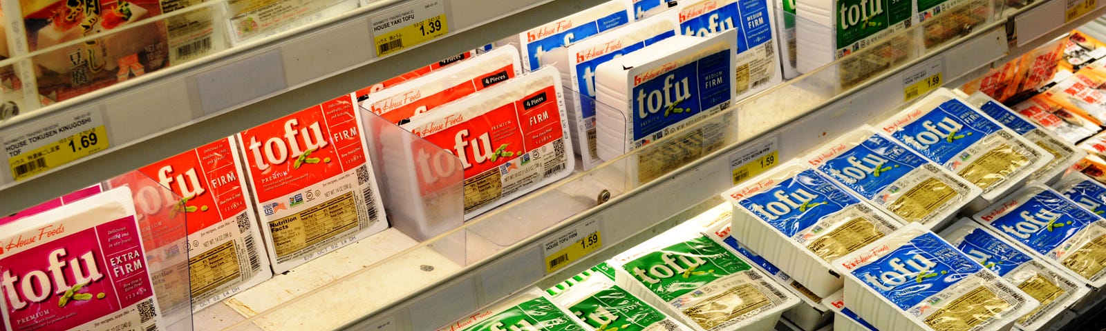 A grocery store refrigerated shelf stocked with many tubs of tofu — firm, extra-firm, and silken in different colors