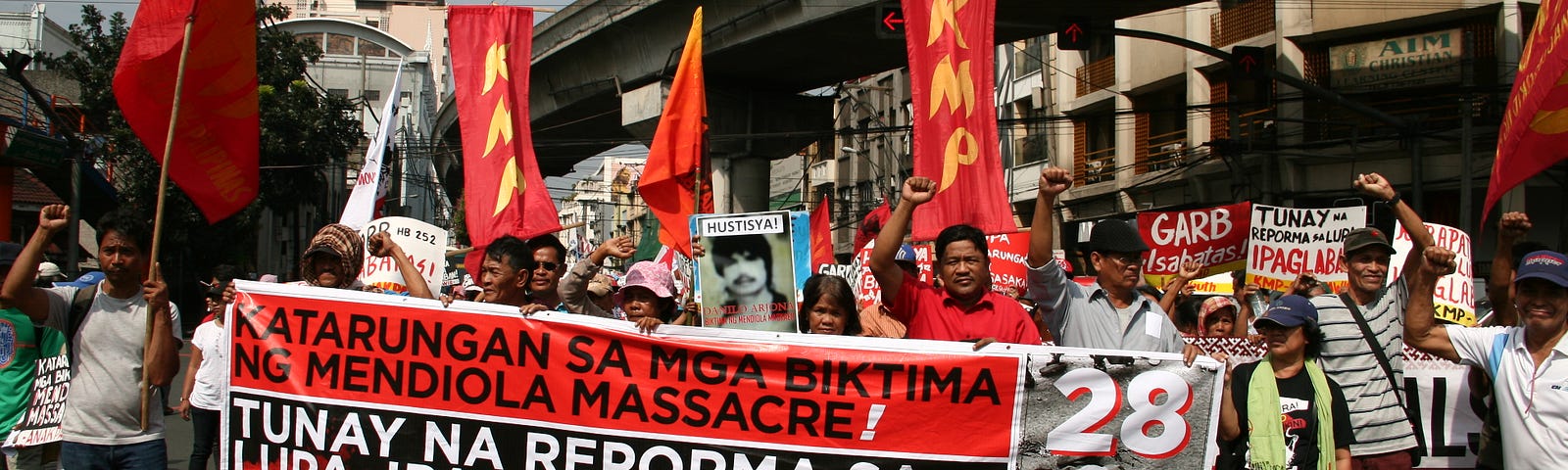 A crowd of protesters holding vertical red flags and carrying a large banner with Tagalog writing.