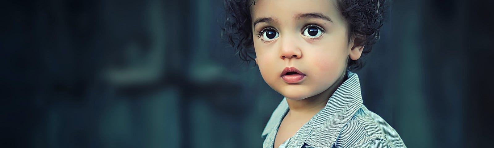 Toddler with brown hair staring at the camera. Dark blue wall in the background. Toddler is wearing a blue shirt.