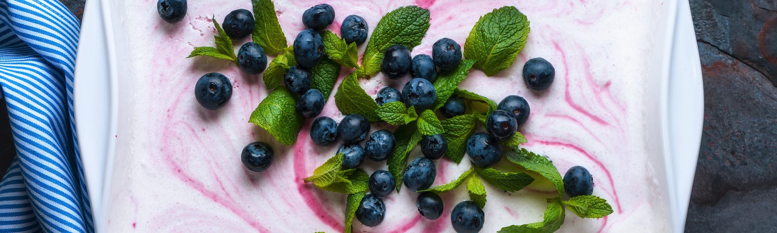 Overhead view of a rectangular ceramic dish with white ice cream with a red swirl & garnished with blueberries + mint leaves.