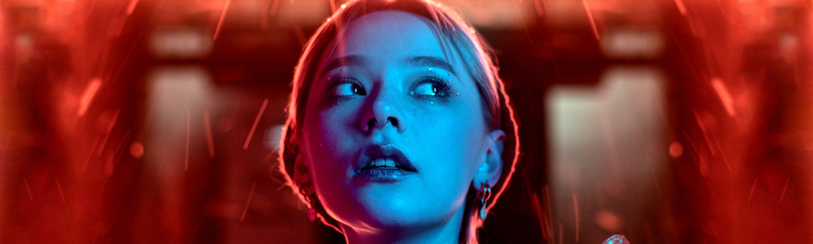 A woman is standing below bright red neon lights. She has a short blond hair, is wearing a black shirt and is holding a shoe.