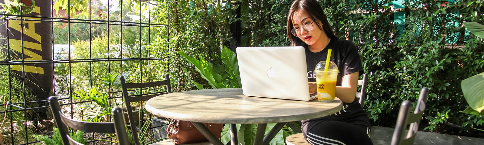 Woman sitting at a table, working on her laptop. She is on a lawn, surrounded by plants and small trees in an indoor/outdoor hybrid setting. She has a yellow beverage next to her laptop.