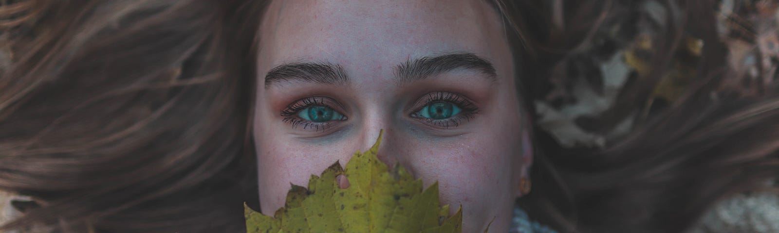 Person looks at camera, while lying in autumn leaves, covering mouth with a yellow leaf.