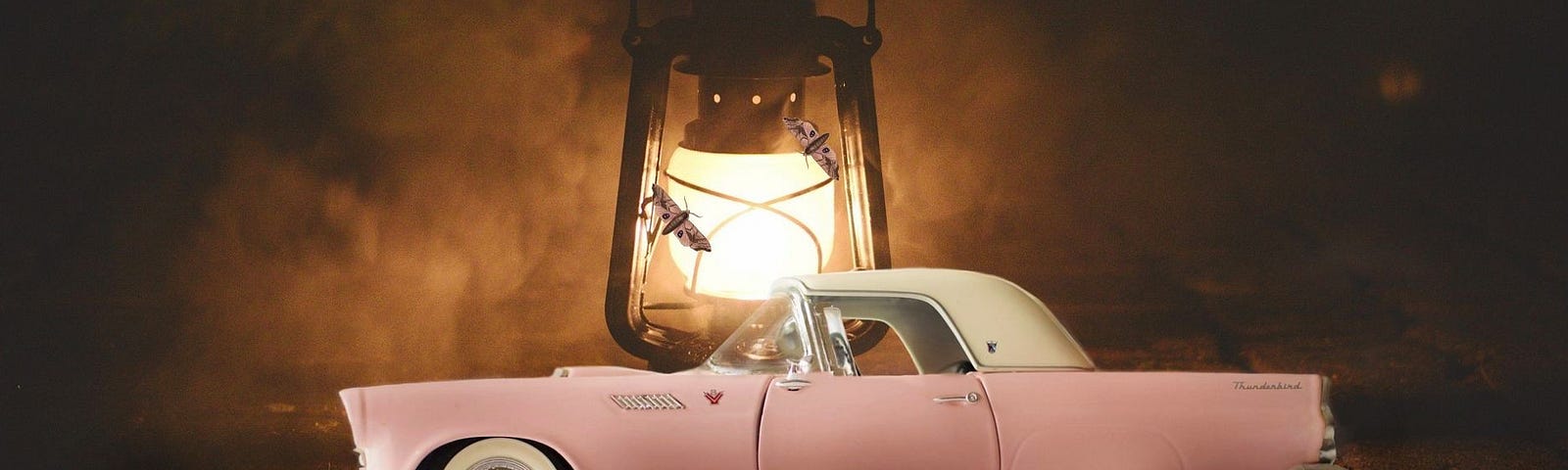 A pink and white vintage two-door coupe parked in front of a large hurricane lantern with moths fluttering around it and set against a dark, foggy background.