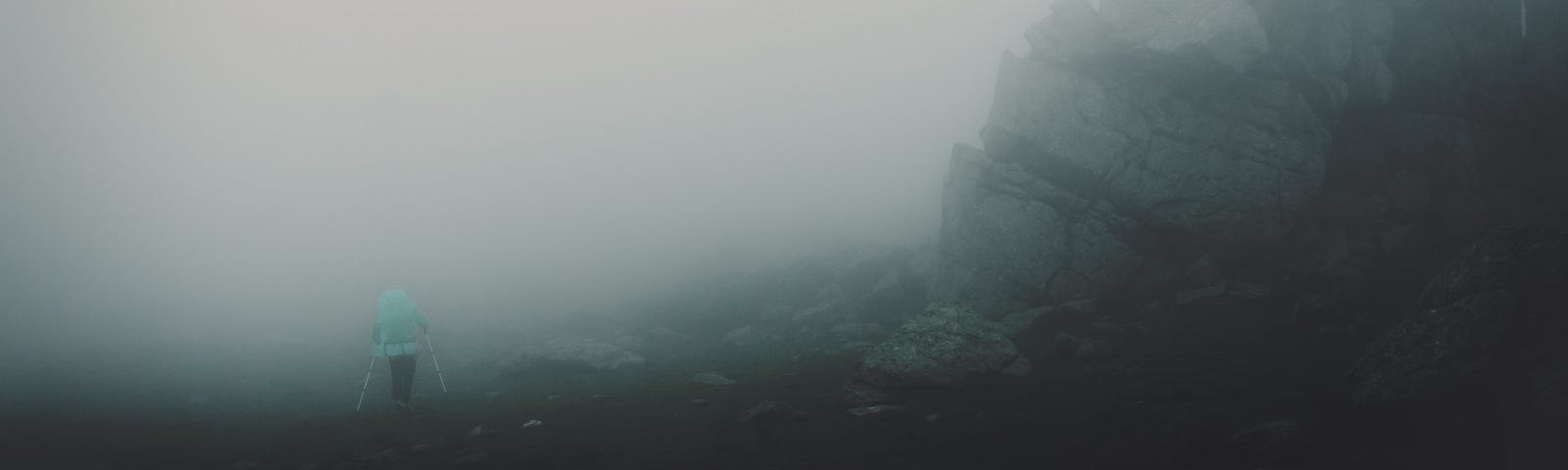 A person hikes along the base of a hill. It’s foggy.