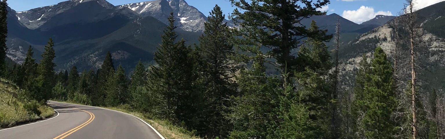 A road curving off into trees with mountains in the background and a blue sky with a few puffy clouds