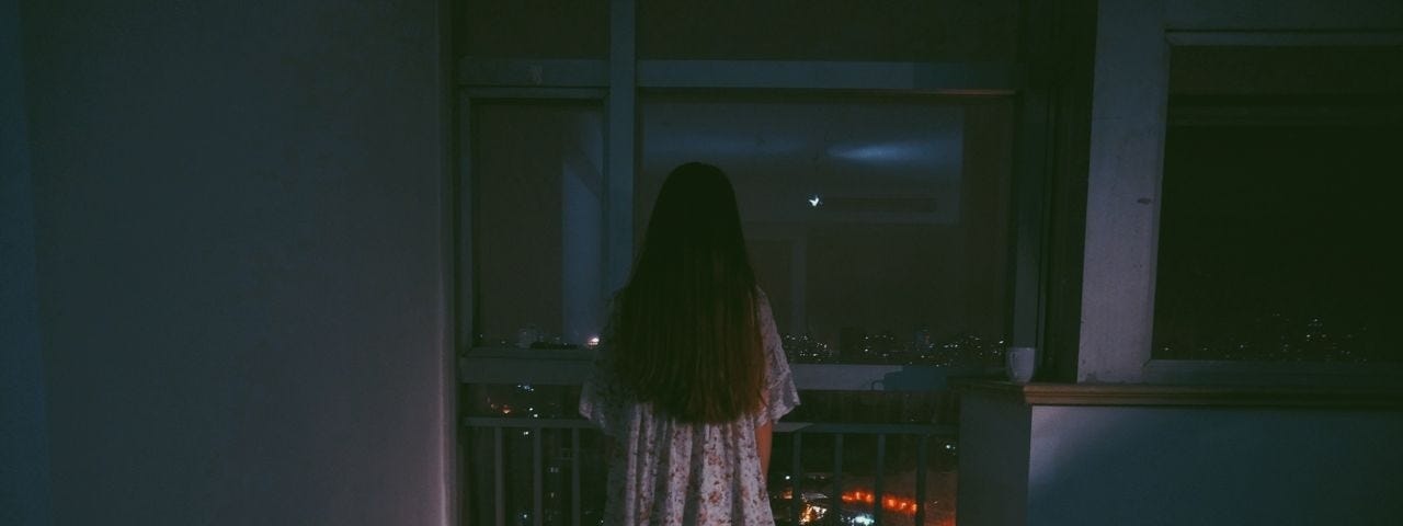 An ominous photo of a girl with long hair looking out a window at night.