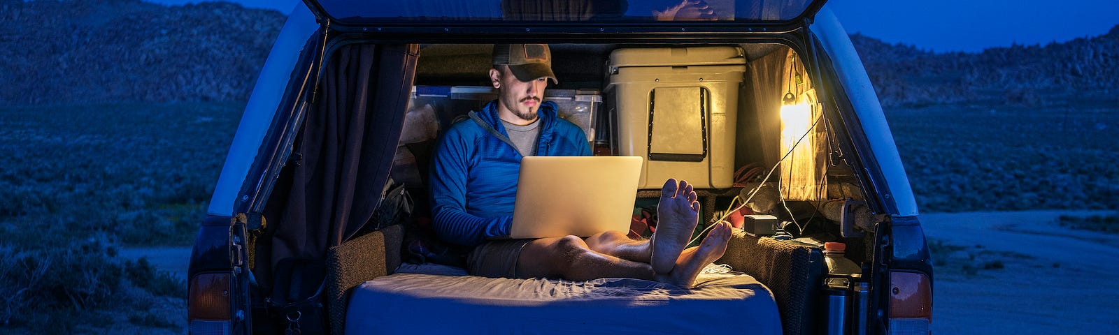A person working on a laptop in the back of a covered pickup truck at night, sitting on a mattress.