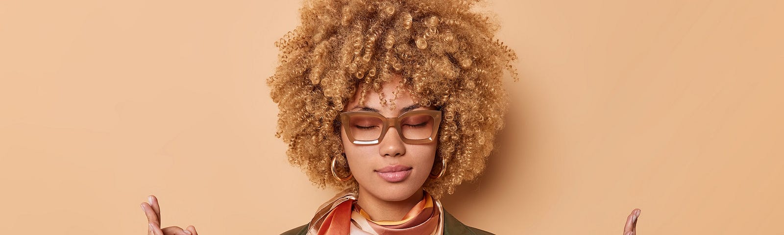 Calm relaxed curly young woman gathers patience makes mudra sign breathes deeply keeps eyes closed wears leather shirt scarf tied around neck reaches nirvana isolated over beige background.