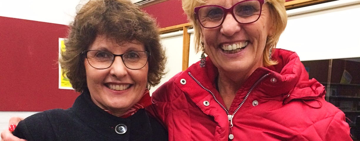 Two women smiling at the camera, one with dark hair and a black coat, and the other fair-haired and wearing a red coat while holding a book, Reversed: A Memoir by Lois Letchford.