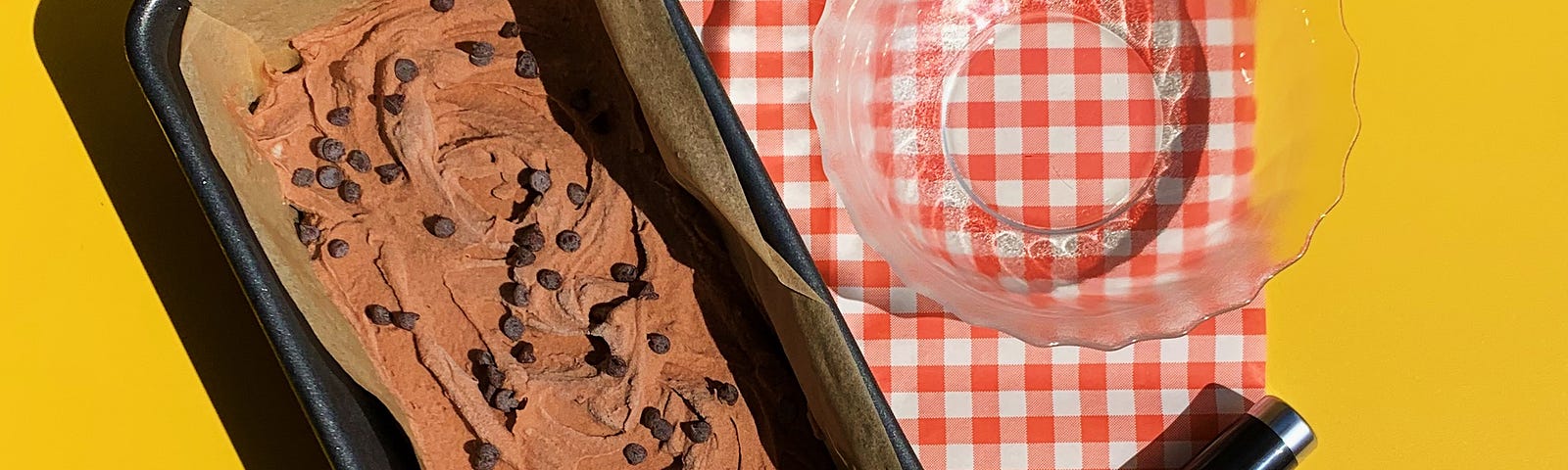 A loaf pan lined with parchment paper and filled with chocolate chocolate chip ice cream next to an ice cream scoop & a dish.