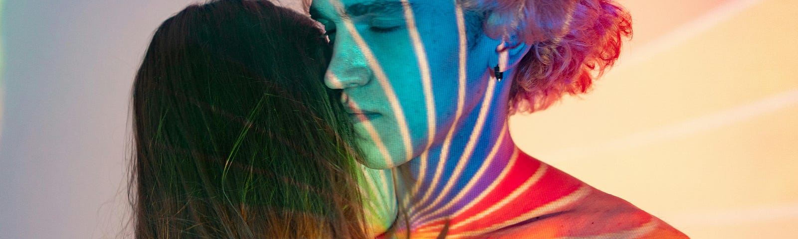 Two men embrace in a hug, one man has blond hair and his eyes are closed. The other has long, dark hair, and his face is out of view. They both have swirling, rainbow colors projected onto their faces and shoulders.