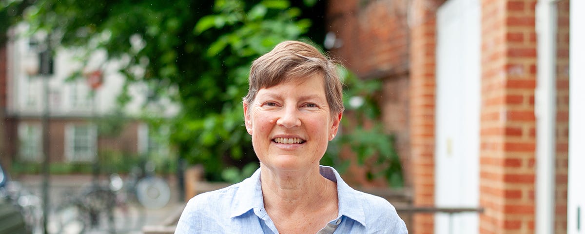 A photo of Joanna Elson, a woman with short brown hair who smiles at the camera. She wears a pale blue shirt open at the collar and stands outside for the photo.