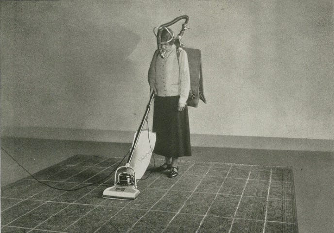 A person wearing a gas mask that connects to a bag on their back uses an upright vacuum on a carpet with a rectangular grid.