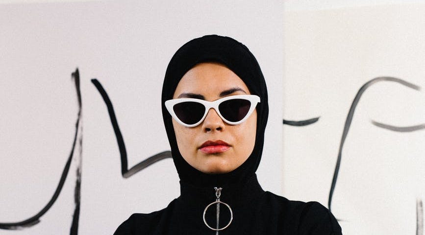 The image shows a woman wearing a black hijab, tucked into a O-ring zip sweatshirt, and white cat-eye sunglasses.