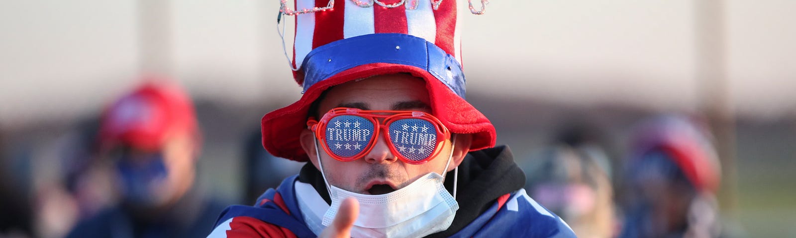 A Trump supporter at an election eve rally wears a US flag hat with lights spelling Trump and a MAGA flag around his neck