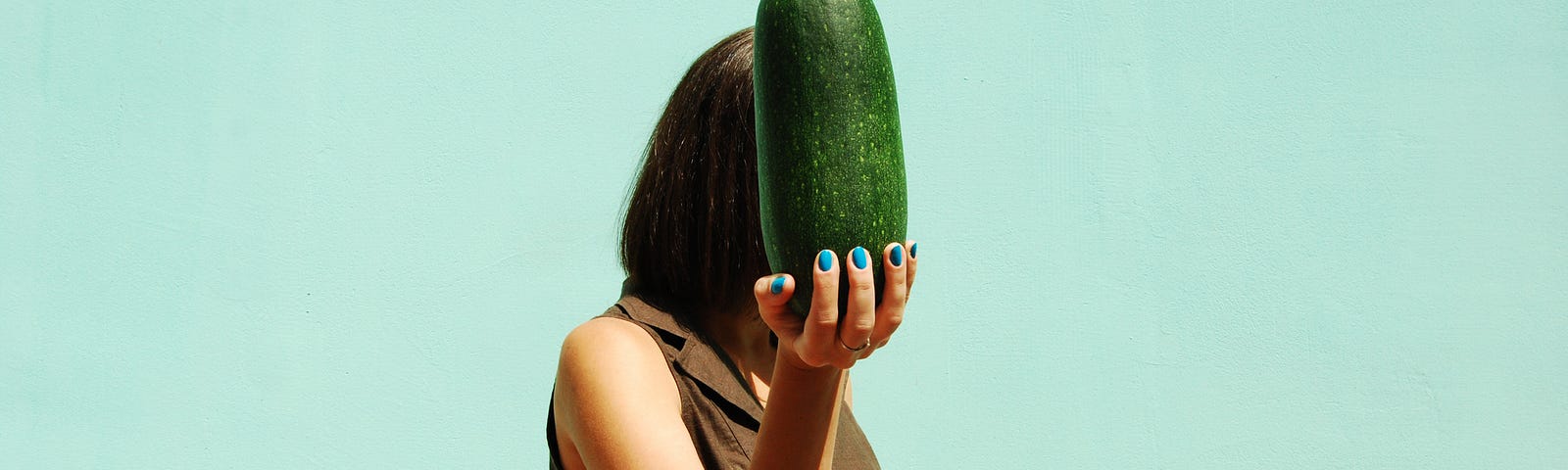 A photo of a woman holding a large cucumber that covers her face, against a mint/teal background.