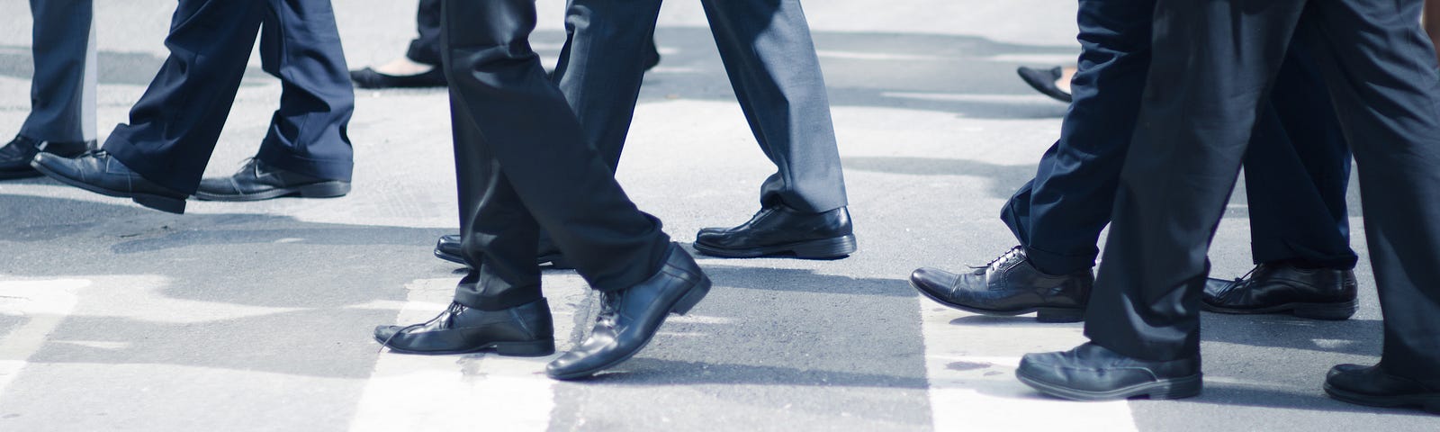 Group of businessmen in similar dark navy suits crossing a city street. You can just barely see the feet of two women.