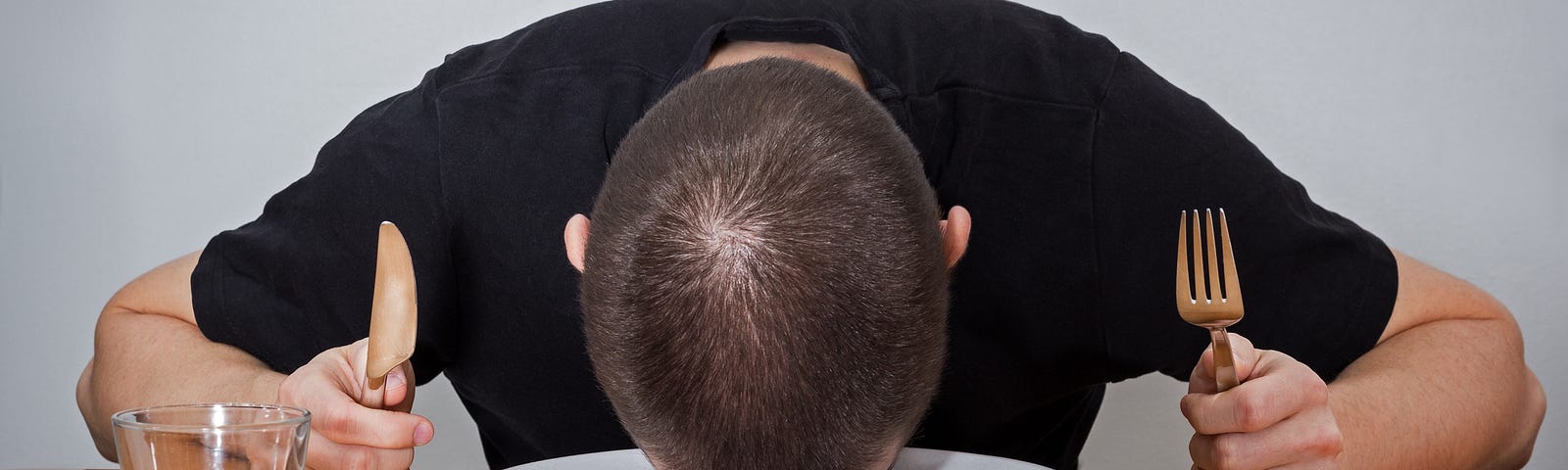 A person holding a fork and knife at a dinner table, resting their head on an empty plate.