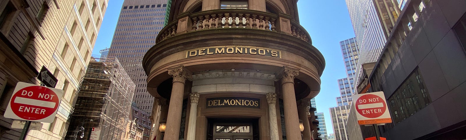 Wide-angle shot of the entrance to Delmonico’s, a skinny building flanked on either side by 1-way streets and skyscrapers.