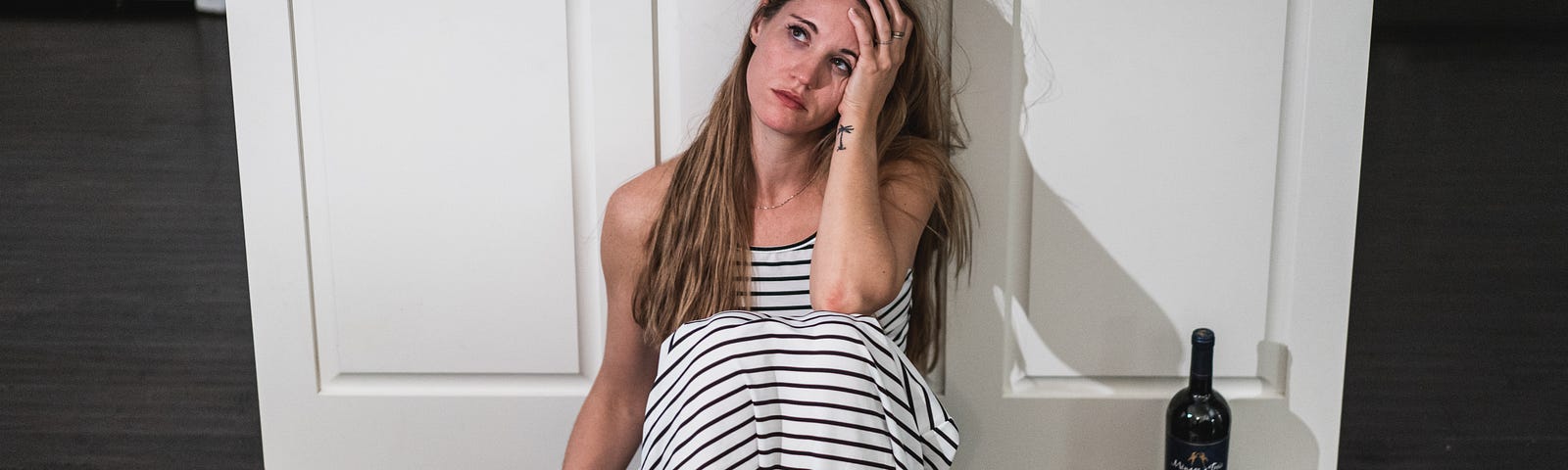 Girl in a white and black horizontally striped dress sitting on the floor in a kitchen with her back against the cabinets -glass of wine in hand with the bottle next to her on the floor