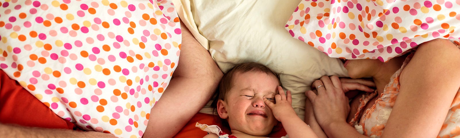 Exhausted parents covering their faces with pillows as baby cries between them.