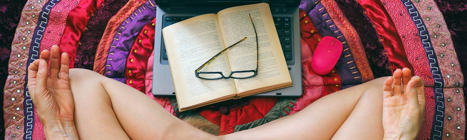 A photo of a woman sitting cross-legged, meditating, on the floor. A laptop with an open book and glasses is in front of her.