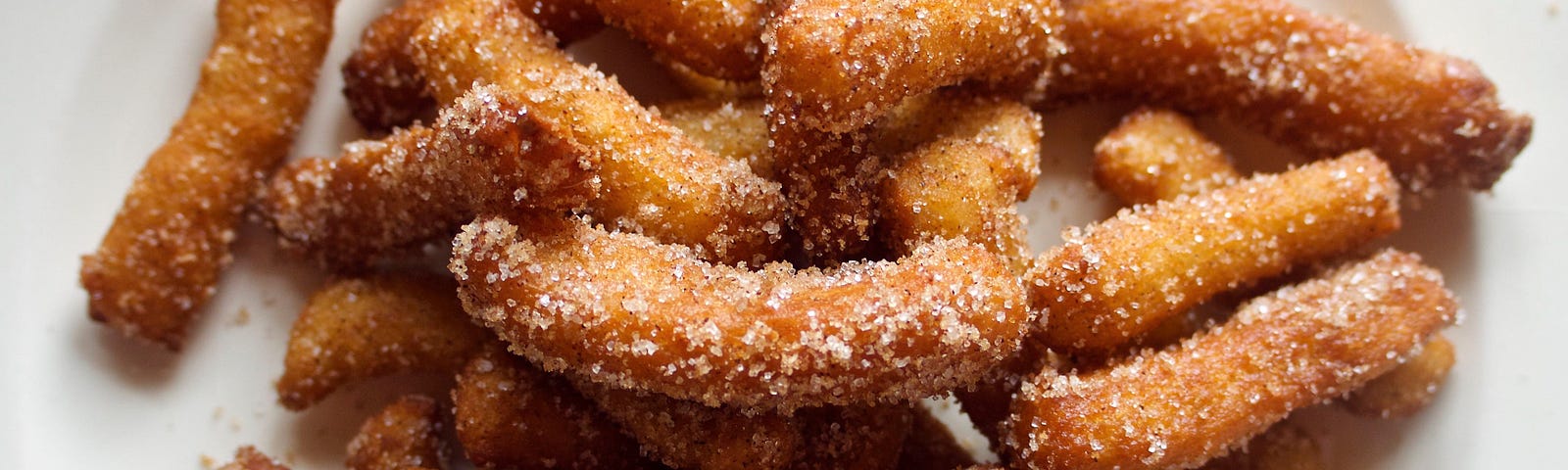 A photo of a plate piled high with homemade golden fried churros, tube-shaped dough that’s dusted in sugar and cinnamon