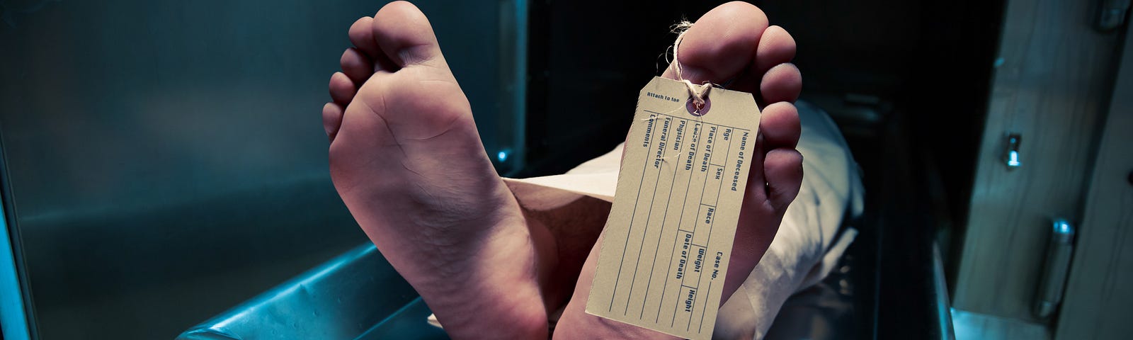 Photo of a body with a toe tag. It’s on a table that is partially inside a cupboard, like you see in a morgue.