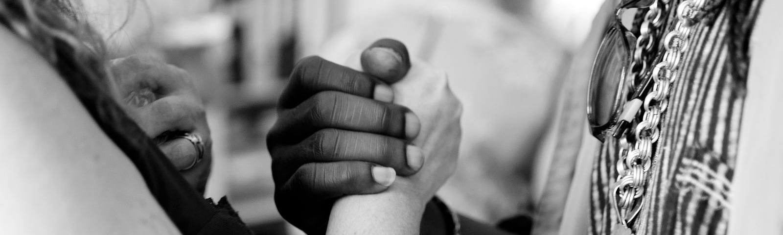 Extreme close-up view in black and white of a woman’s white hand interlocked with a man’s black hand, her arm bare, his chest showing heavy jewelry and sunglasses hung there, with a soft-focus view of others in the background.