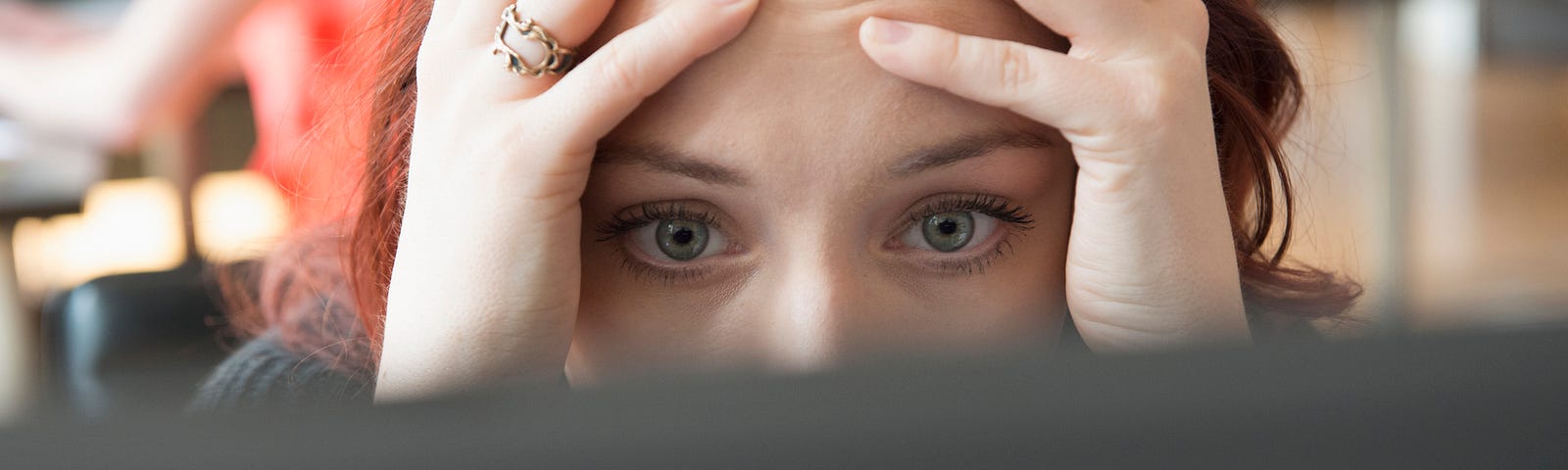 A photo of a stressed woman with her hands on the sides of her face, looking at her computer screen.