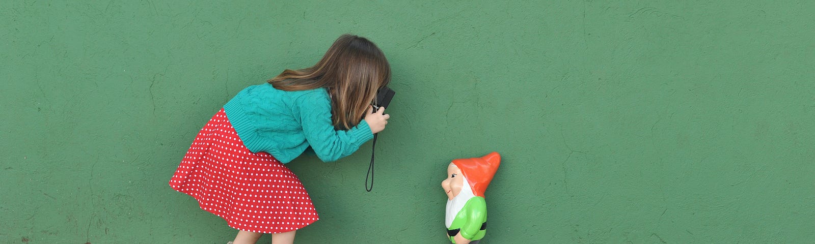 A little girl in a green sweater and polka dot skirt taking a photo of a happy gnome.