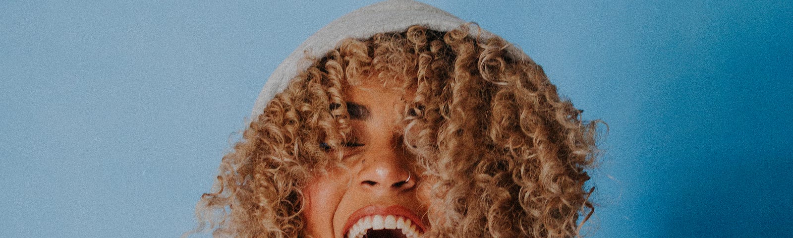 Young golden-skinned woman with tight dark blond curls and a wide smile with both hands cupped at her neckline, wearing a gray hoodie, hood up, against a blue background.