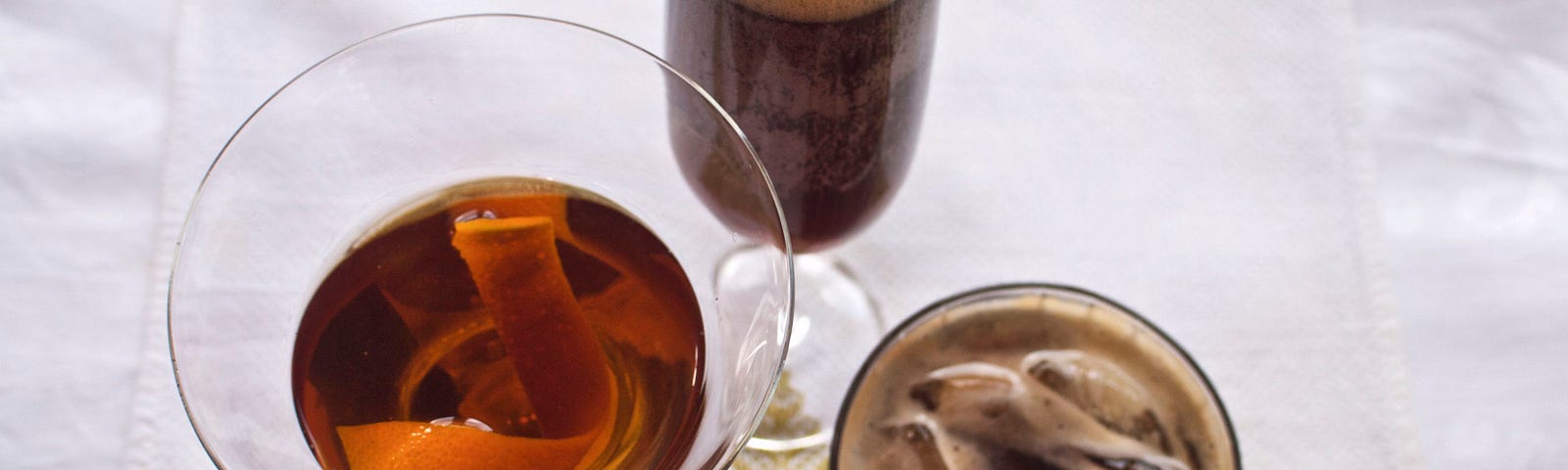 A trio of cocktails photographed from above: one is in a martini glass w orange peel, one tall w foam, one dark on the rocks