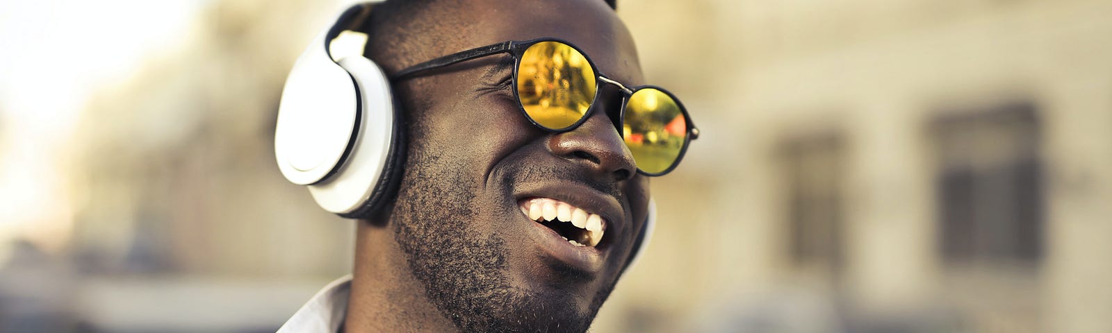 Photograph of black man wearing white headphones and white shirt, smiling with mouth open and wearing yellow tinted sunglasses. Blurred buildings and cars in background. Feeling of happiness.