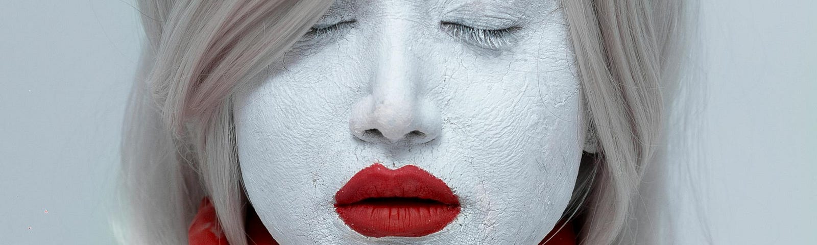 A silver-haired woman wearing white face and body paint poses with her eyes closed. Her lips are colored red, as are her forearms. Her hands are placed around her neck in an artful pose. The backdrop is grey.