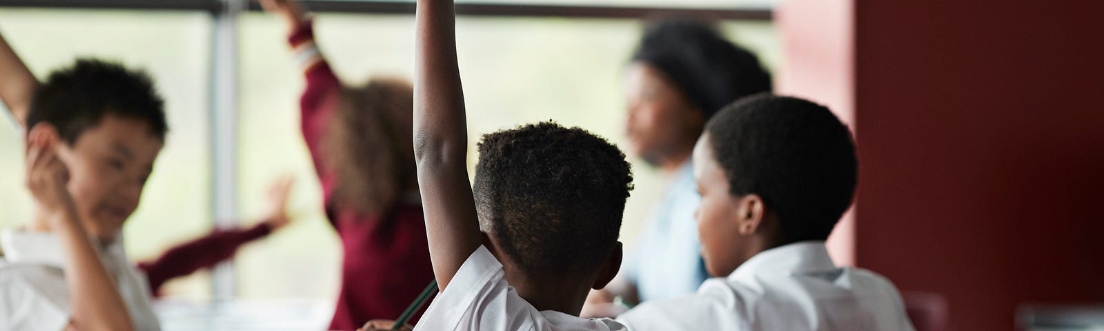 A photo of a young black boy with his hand raised at his desk.