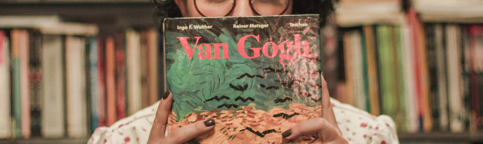 A woman is standing in front of shelves of books, holding a “Van Gogh” book. She is hiding her mouth behind the book, with her eyes and glasses peeking out over the top.
