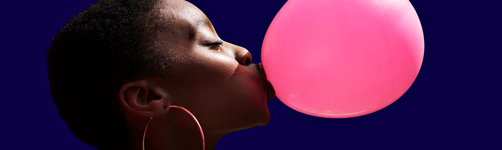 A photo of a young black woman wearing hoop earrings blowing a bubble with bubblegum.