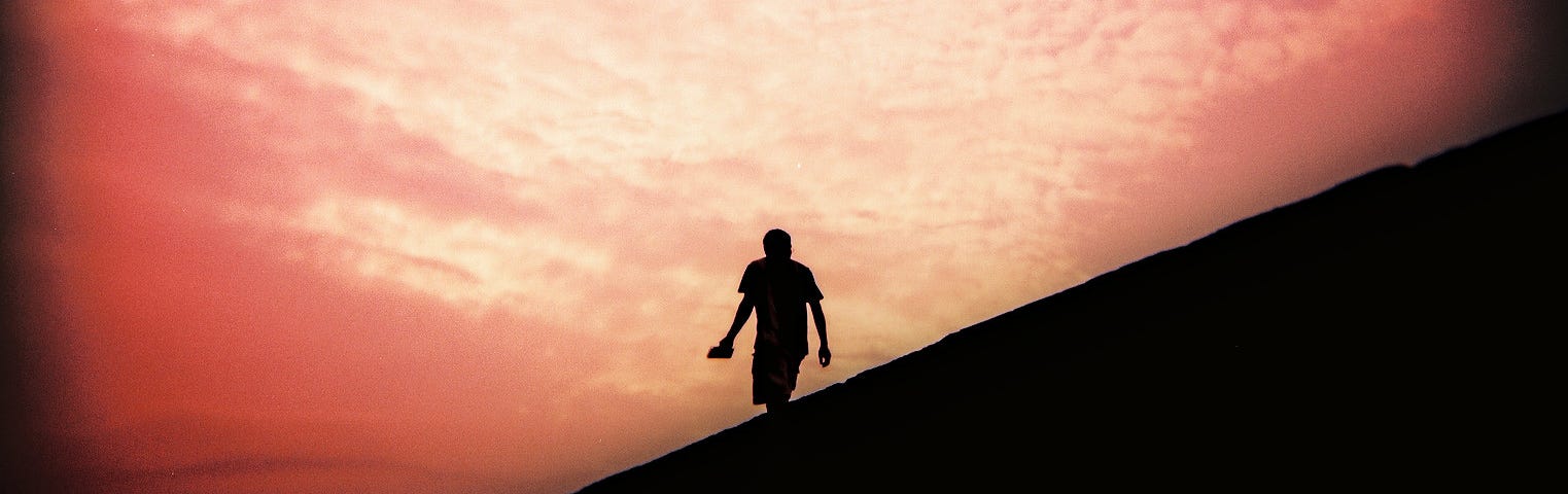 Photo of the dark silhouette of a person walking in front of a pink sunset.