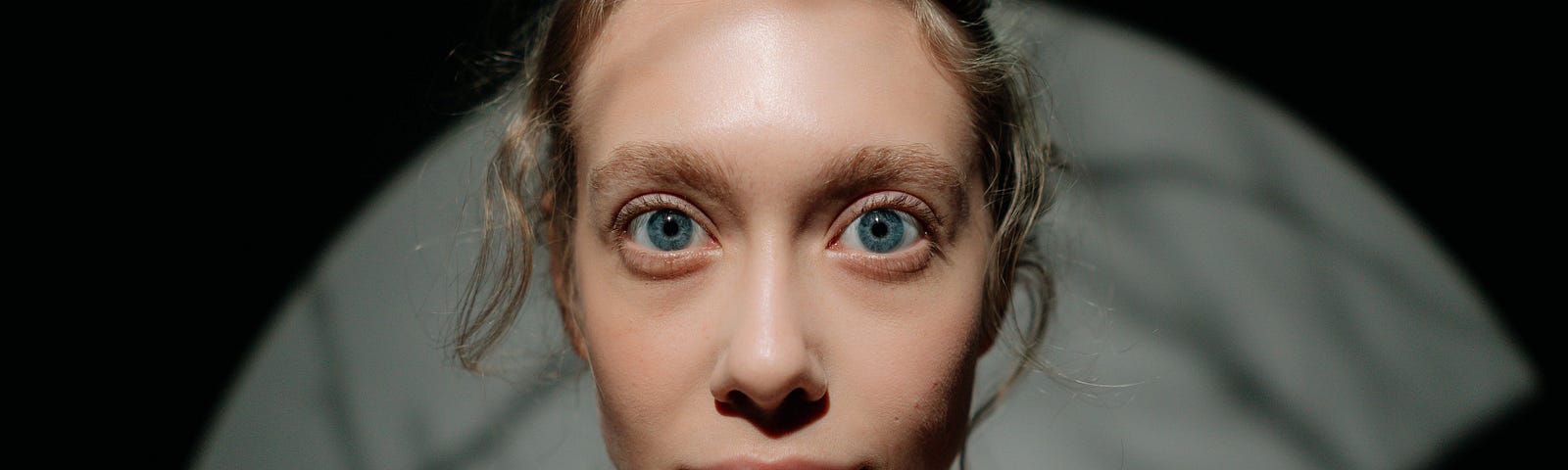 An image of a blond-haired woman with bright blue eyes staring at the camera with a slightly eccentric expression, almost as though she’s possessed by something. There are shadows in the background, on the right side of the image is a shadow of a dark hand reaching out to touch her.