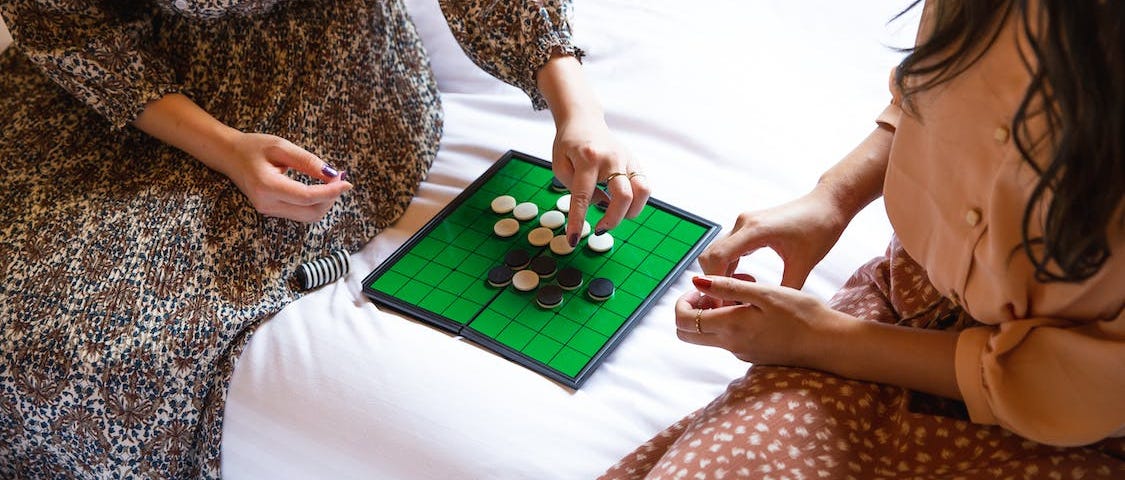 Two women, faces unseen, playing Othello on a green board with black & white reversible disks.