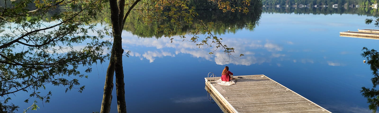 clear blue skies, a calm peaceful lake showing a reflection of the clouds and trees, and a woman sitting on the dock enjoying the view