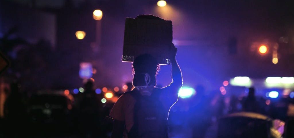 A dark nighttime photo of a protestor holding up a sign with police lights in the background.