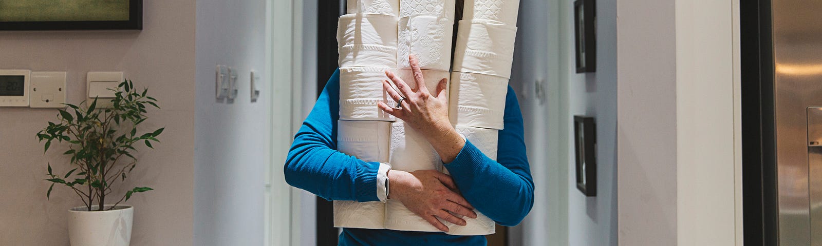 A photo of a person holding a tower of toilet paper rolls as they walk down the hallway.