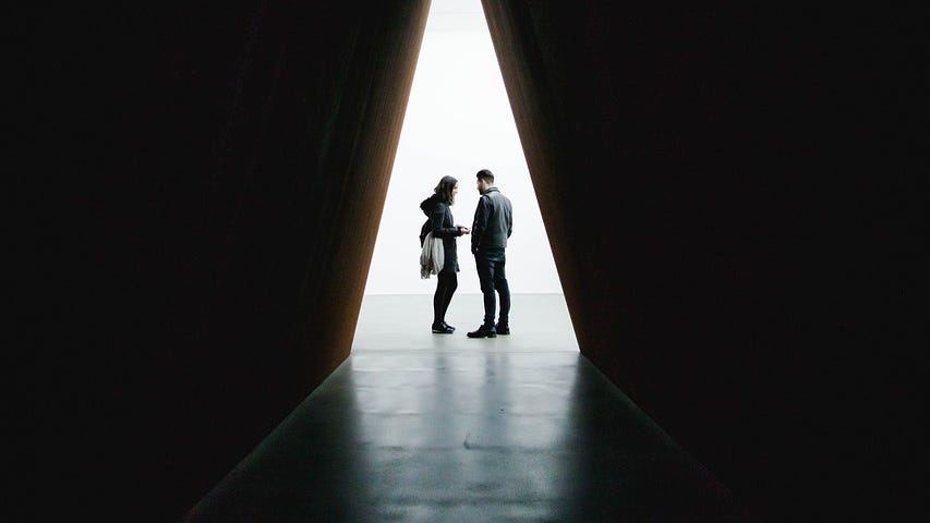 A man and woman standing in profile and conversing in the triangular-shaped opening between two very tall black walls.