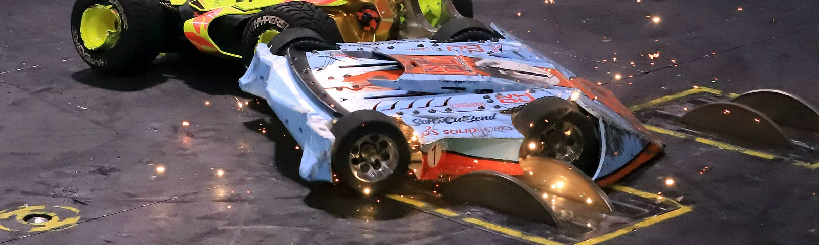 A still image from BattleBots 2022 Championship: HyperShock vs P1. HyperShock sports florescent yellow plastic armor and a duel toothed disk spinner in front. P1 is has a blue and orange paint job reminiscent of a formula 1 race car. It feature a flipper that pivots near it’s front edge. In the image, HyperShock is ramming P1 into two kill saws that have popped up from two slits in the floor.