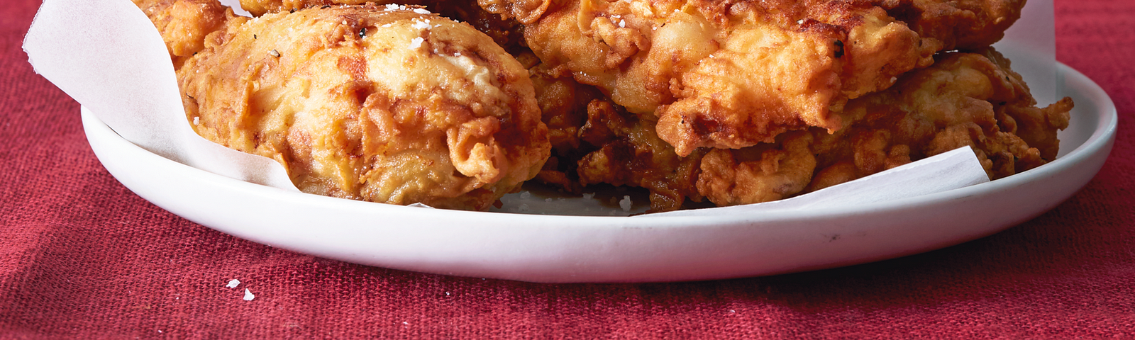 A white dish with a heap of fried chicken parts, on a red tablecloth with a red background.