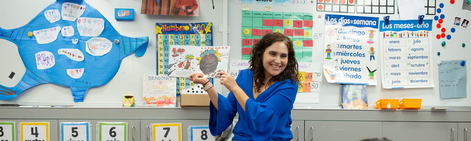 A female teacher shows an open book to young students sitting on the floor of a classroom.